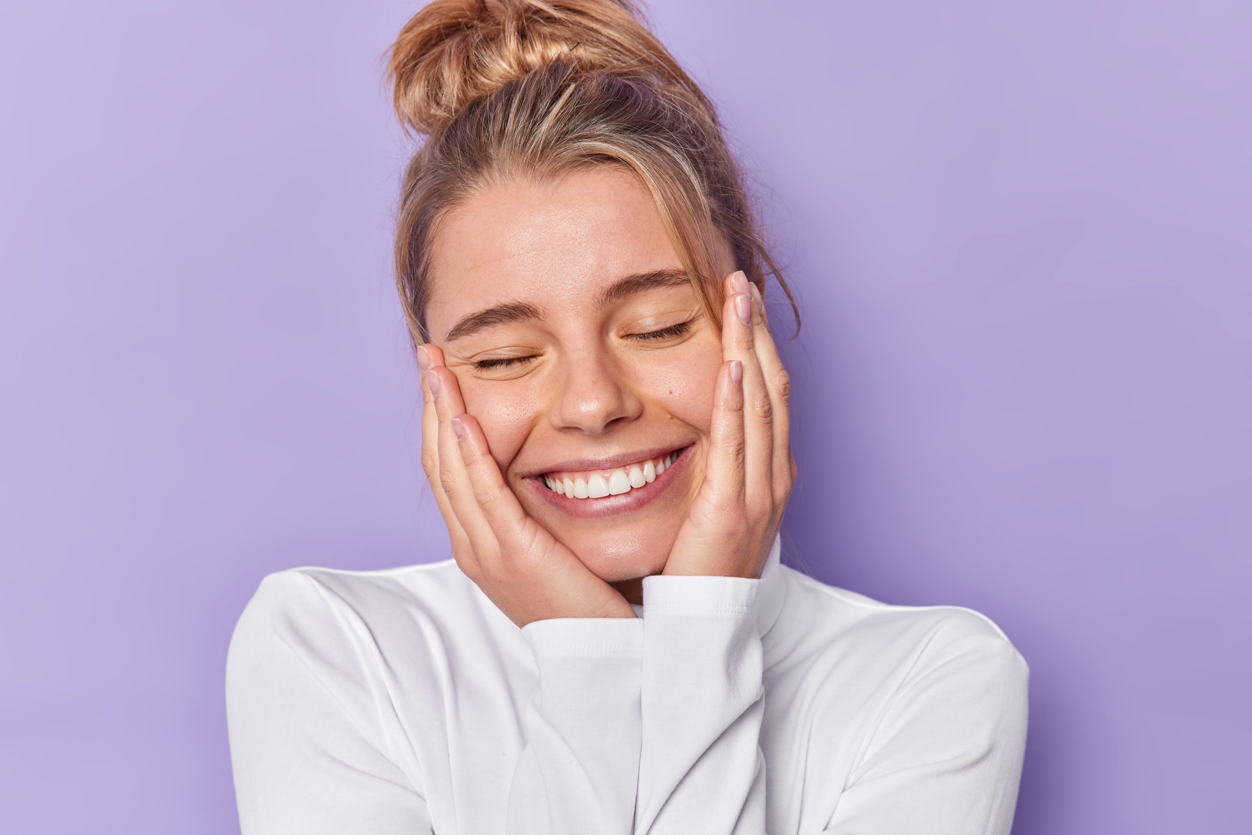 woman-keeps-eyes-closed-smiles-broadly-has-combed-hair-wears-white-casual-jumper-isolated-purple-studio-positive-young-female-model-glad-hear-compliment-scaled.jpg